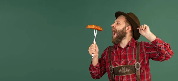 Retrato de hombre barbudo en traje tradicional bávaro con salchichas divirtiéndose aisladas sobre fondo verde. Oktoberfest, festival, concepto de tradiciones — Foto de Stock