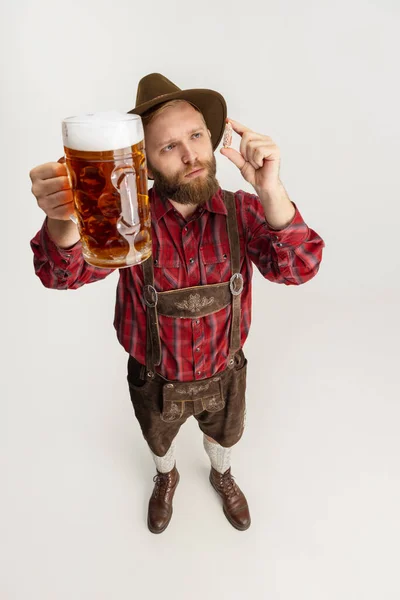 Retrato cómico del hombre barbudo con sombrero y traje bávaro tradicional que sostiene una taza enorme, un vaso de cerveza espumosa ligera y aislado sobre un fondo blanco. Oktoberfest — Foto de Stock