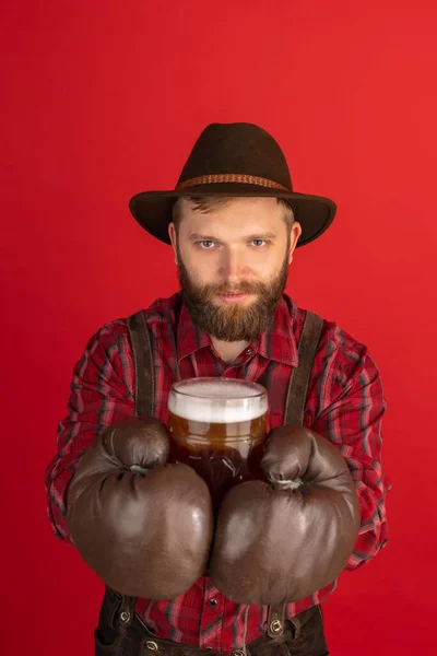 Portrait comique de l'homme barbu, serveur en chapeau et costume bavarois traditionnel isolé sur fond de studio rouge. Oktoberfest, fest, concept vacances — Photo