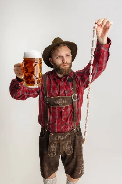 Комический портрет бородатого человека в шляпе и традиционном баварском костюме, держащего огромную кружку, стакан светлого пенного пива и изолированного на белом фоне. Октоберфест — стоковое фото
