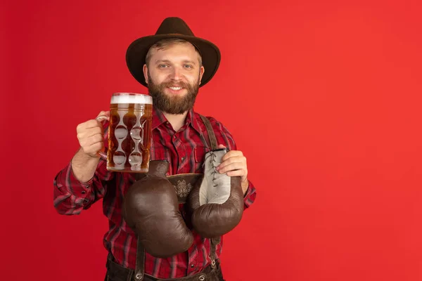 Comic portrait of bearded man, waiter in hat and traditional Bavarian costume isolated over red studio background. Oktoberfest, fest, holidays concept — Stock Photo, Image