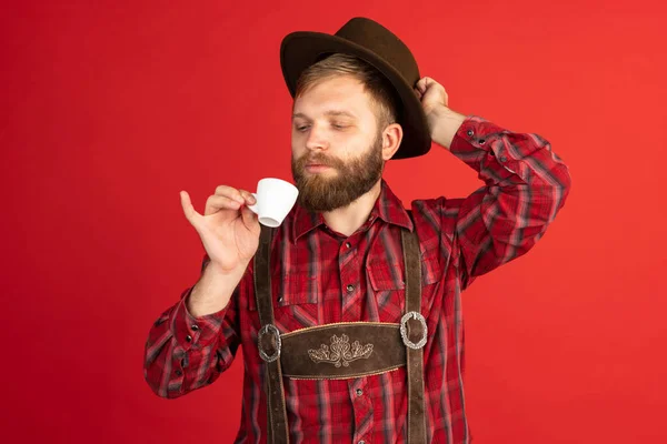 Retrato cómico de hombre barbudo, camarero con sombrero y traje bávaro tradicional aislado sobre fondo rojo del estudio. Oktoberfest, fest, concepto de vacaciones — Foto de Stock