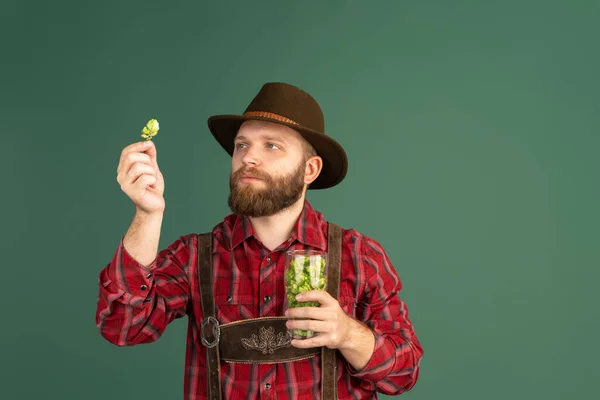 Ritratto di uomo barbuto, birraio in costume tradizionale bavarese con bicchieri di birra pieni di selvaggio caldo isolato su sfondo verde. — Foto Stock