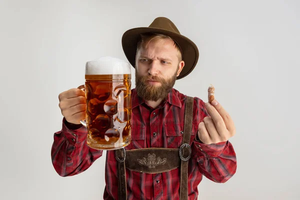 Half-length portrait of bearded man in hat and traditional Bavarian costume holding huge mug, glass of light frothy beer isolated over white background. Flyer — Stock Photo, Image