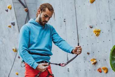 Güvenli tırmanış. Genç adam profesyonel kaya tırmanışçısı spor malzemelerini kontrol ediyor güneşli bir günde, açık havada eğitim merkezine tırmanmadan önce..