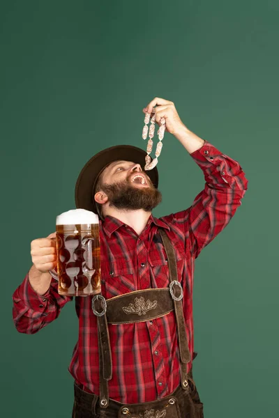 Retrato de hombre barbudo en traje tradicional bávaro con salchichas divirtiéndose aisladas sobre fondo verde. Oktoberfest, festival, concepto de tradiciones —  Fotos de Stock