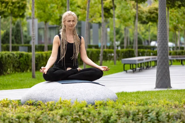 Mulher bonita nova com cabelo loiro fazendo exercício de ioga no parque público verde na manhã de verão, ao ar livre. Estilo de vida saudável, conceito de saúde mental. — Fotografia de Stock