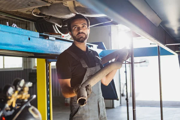 Ein junger Mann, Automechaniker, Angestellter in Latzhosen, der in der Garage einen Autoservice macht, drinnen. Arbeitsbegriff, Beruf, Geschäft, Pflege, Job — Stockfoto