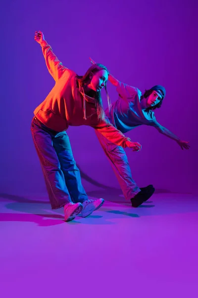 Twee dansers, jonge man en vrouw dansen hip-hop in casual sport jeugd kleding op gradiënt paars roze achtergrond in danszaal in neon licht. — Stockfoto