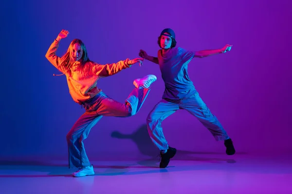 Joven hombre y mujer bailando hip-hop en deportes informales ropa juvenil sobre fondo rosa púrpura degradado en el salón de baile en luz de neón. — Foto de Stock