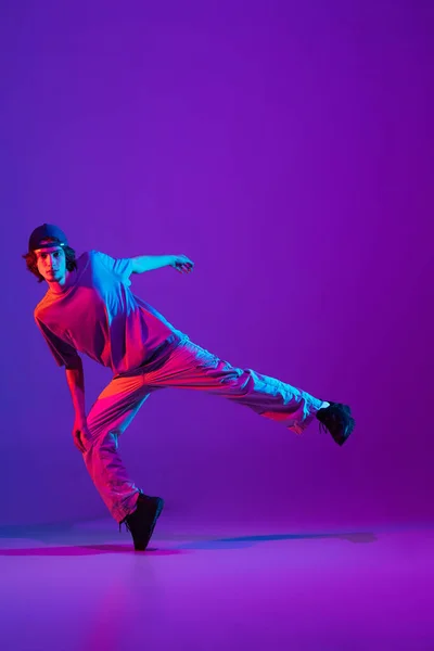 Stijlvolle sportieve jongen dansend hip-hop in stijlvolle kleding op kleurrijke achtergrond in de danszaal in neon licht. Jeugdcultuur, beweging, stijl en mode, actie. — Stockfoto