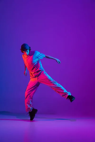 Mladý stylový muž, break dance, hip-hop tanečník cvičit sólo v ležérní oblečení izolované přes fialové pozadí v neonově růžové světlo v tanečním sále — Stock fotografie