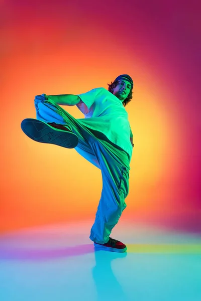 Conceptueel portret van jonge man, hip-hop danser in stijlvolle kleding in actie geïsoleerd op kleurrijke achtergrond in de danszaal in neon licht. — Stockfoto