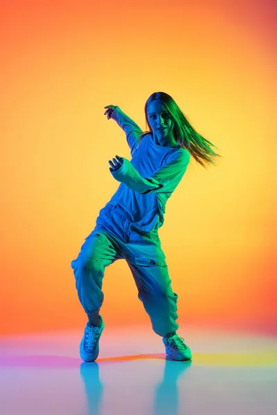Jong sportief meisje dansend hip-hop in stijlvolle kleding op kleurrijke achtergrond in de danszaal in neon licht. Jeugdcultuur, beweging, stijl en mode, actie. — Stockfoto