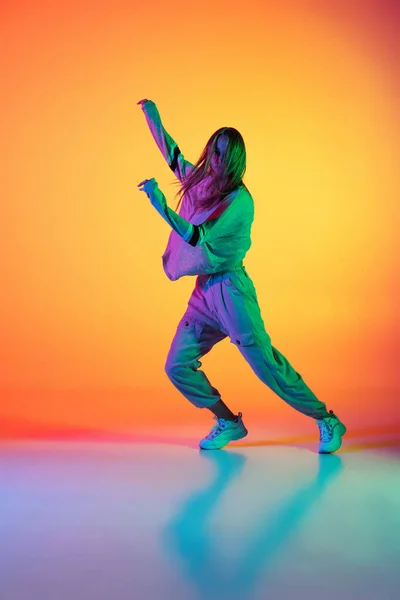 Una chica atlética caucásica bailando hip-hop con ropa elegante sobre un fondo colorido en el salón de baile con luz de neón. Cultura juvenil, movimiento, estilo y moda, acción. — Foto de Stock