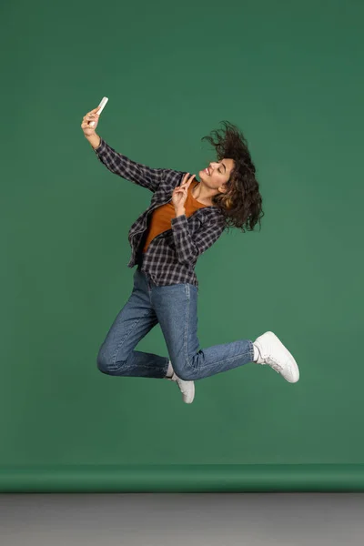 Retrato de una hermosa chica latina saltando y usando un teléfono aislado sobre un fondo de estudio de color verde vintage. Concepto de emociones humanas — Foto de Stock