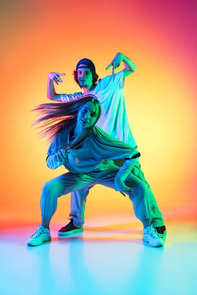 Jovens dançarinos de hip-hop, elegante menina emotiva e menino em ação e movimento em esportes casuais roupas de jovens no gradiente de fundo multi colorido no salão de dança em luz de néon. — Fotografia de Stock