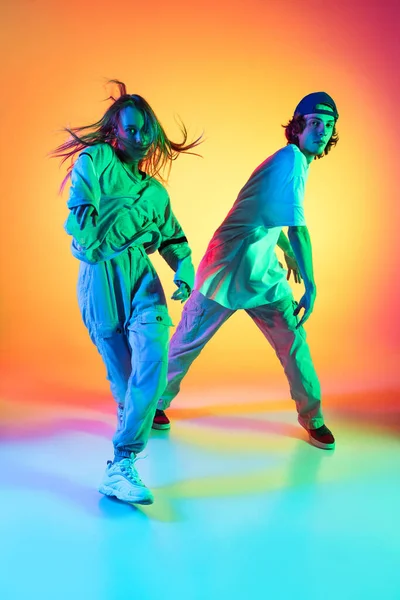 Twee dansers, jong meisje en jongen dansen hip-hop in casual sport jeugd kleding op gradiënt multi gekleurde achtergrond in danszaal in neon licht. — Stockfoto