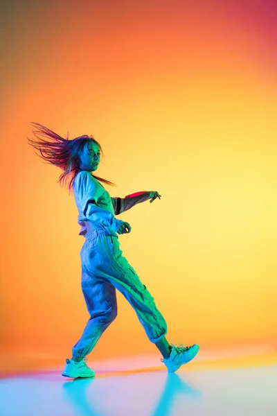 Młoda sportowa dziewczyna tańcząca hip-hop w stylowych ubraniach na kolorowym tle w sali tanecznej w neonowym świetle. Kultura młodzieżowa, ruch, styl i moda, akcja. — Zdjęcie stockowe