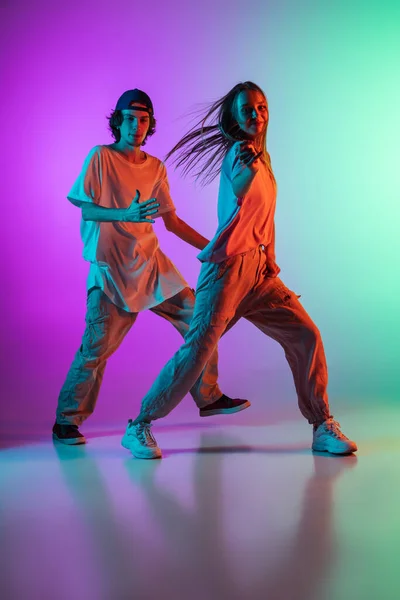 Mladí hip-hop tanečníci, stylové emotivní dívka a chlapec v akci a pohybu v příležitostných sportovních mládežnických oblečení na gradient vícebarevné pozadí v tanečním sále v neonovém světle. — Stock fotografie