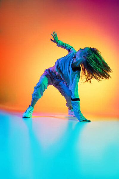 Sportive flexible girl dancing hip-hop in stylish clothes on colorful background at dance hall in neon light. Youth culture, movement, style and fashion, action.