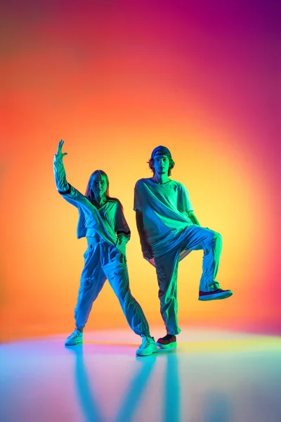 Jonge dansers, stijlvol emotioneel meisje en jongen dansen hip-hop in casual sport jeugd kleding op gradiënt multi gekleurde achtergrond in danszaal in neon licht. — Stockfoto