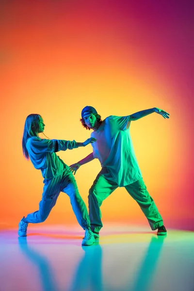 Jonge hiphop dansers, stijlvol emotioneel meisje en jongen in actie en beweging in casual sport jeugd kleding op gradiënt multi gekleurde achtergrond in danszaal in neon licht. — Stockfoto