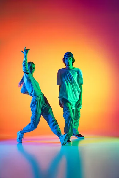 Twee dansers, jong meisje en jongen dansen hip-hop in casual sport jeugd kleding op gradiënt multi gekleurde achtergrond in danszaal in neon licht. — Stockfoto