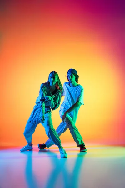 Jonge hiphop dansers, stijlvol emotioneel meisje en jongen in actie en beweging in casual sport jeugd kleding op gradiënt multi gekleurde achtergrond in danszaal in neon licht. — Stockfoto