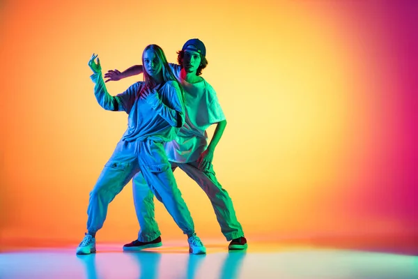 Jovens dançarinos de hip-hop, elegante menina emotiva e menino em ação e movimento em esportes casuais roupas de jovens no gradiente de fundo multi colorido no salão de dança em luz de néon. — Fotografia de Stock