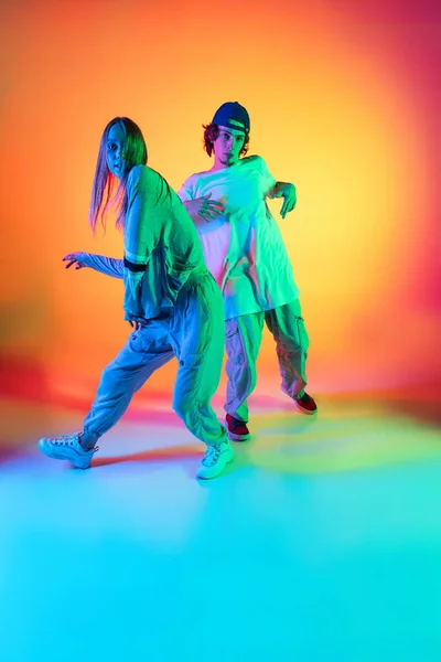 Jonge hiphop dansers, stijlvol emotioneel meisje en jongen in actie en beweging in casual sport jeugd kleding op gradiënt multi gekleurde achtergrond in danszaal in neon licht. — Stockfoto