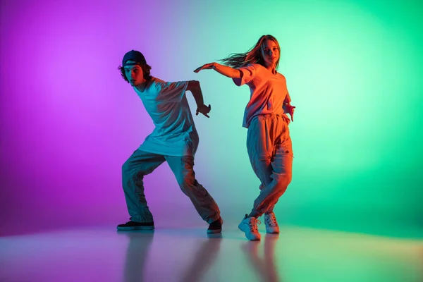 Stijlvolle jonge hip-hop dansers, emotief meisje en jongen in actie en beweging in casual sport jeugd kleding op gradiënt multi gekleurde achtergrond in de danszaal in neon licht. — Stockfoto