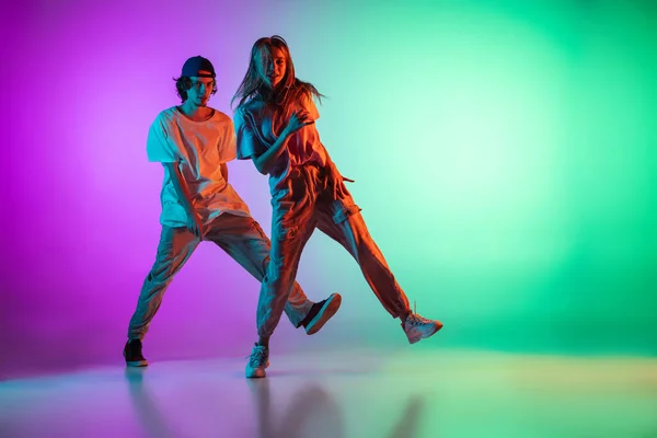 Stijlvolle jonge hip-hop dansers, emotief meisje en jongen in actie en beweging in casual sport jeugd kleding op gradiënt multi gekleurde achtergrond in de danszaal in neon licht. — Stockfoto