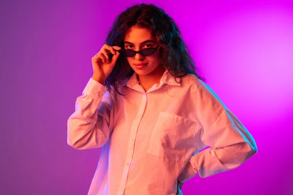 Portret van mooi meisje zetten zonnebril op en kijken naar camera geïsoleerd op paars, lila kleur studio achtergrond in neon licht. — Stockfoto
