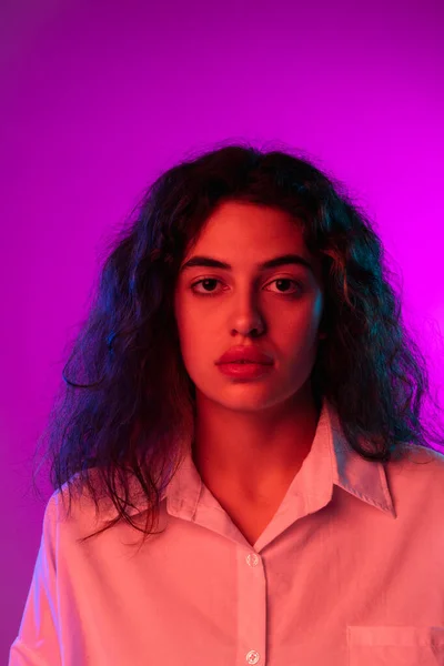 Wanita muda yang menarik melihat kamera terisolasi pada latar belakang studio berwarna ungu dan ungu dalam cahaya neon. Konsep emosi, ekspresi — Stok Foto