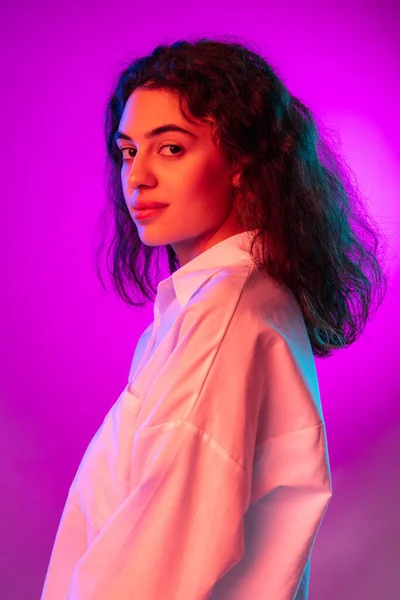 Wanita muda yang menarik melihat kamera terisolasi pada latar belakang studio berwarna ungu dan ungu dalam cahaya neon. Konsep emosi, ekspresi — Stok Foto