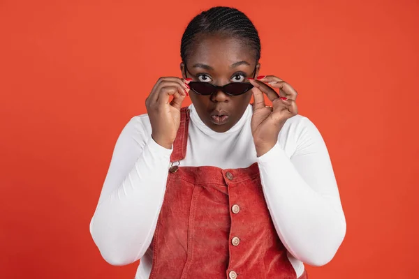Helles Porträt einer überraschten Afrikanerin mit Sonnenbrille, isoliert auf rotem Studiohintergrund. Konzept menschlicher Emotionen, Gesichtsausdruck, natürliche Schönheit, Körperpositiv — Stockfoto