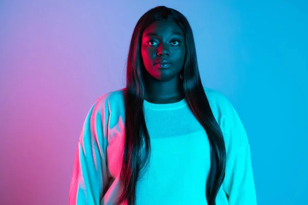 Primer plano de mujer africana encantadora con el pelo largo y liso aislado sobre fondo de estudio azul en luz de neón. Concepto de emociones humanas —  Fotos de Stock