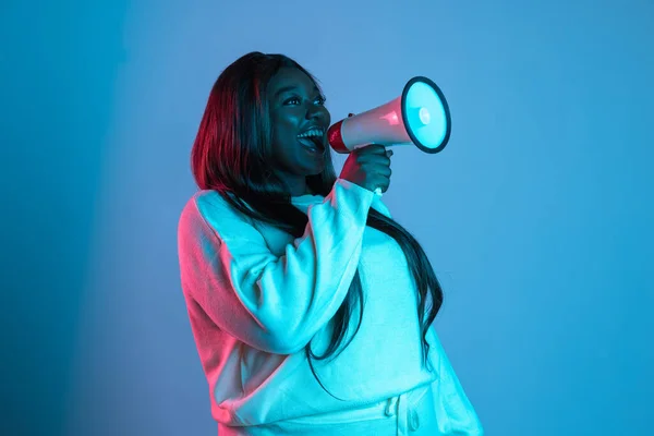 One African beautiful woman with long straight hair shouting at megaphone isolated on blue studio background in neon light. Concept of human emotions — Stock Photo, Image