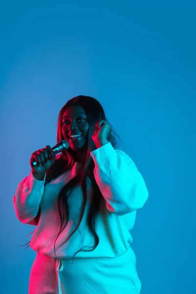 Una mujer hermosa africana alegre con el pelo largo y liso cantando aislado en el fondo del estudio azul en luz de neón. Concepto de emociones humanas —  Fotos de Stock