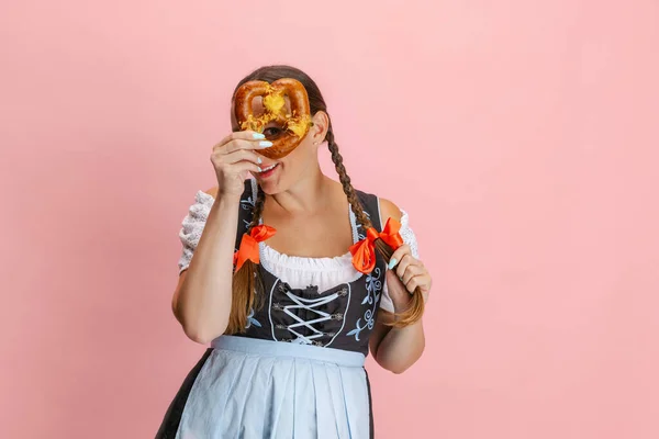 Imádnivaló Oktoberfest nő, pincérnő visel hagyományos bajor vagy német dirndl holding bretzel elszigetelt rózsaszín stúdió háttér. — Stock Fotó