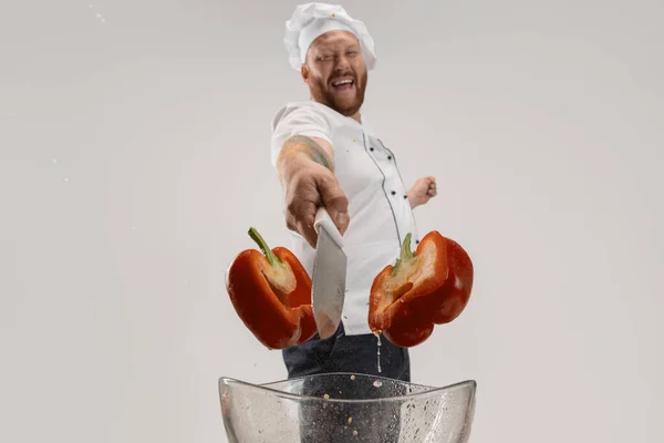 Close-up een hadsome baardman, kok, mannelijke chef-kok in wit uniform snijdt peper geïsoleerd op witte achtergrond. — Stockfoto