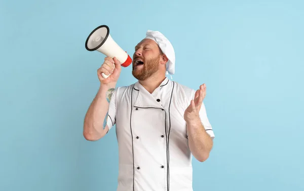 Ritratto comico di uomo barbuto, cuoco, cuoco in uniforme bianca che grida al megafono isolato su sfondo blu. Concetto di lavoro, professione, umorismo. — Foto Stock