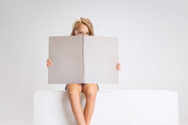 Beyaz stüdyo arka planında izole edilmiş büyük bir kutunun üzerinde oturan günlük kıyafetlerle küçük tatlı bir kızın portresi. Mutlu çocukluk kavramı.