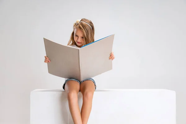 Retrato de una niña linda con ropa casual sentada en una caja grande aislada en un fondo de estudio blanco. Concepto de infancia feliz. —  Fotos de Stock