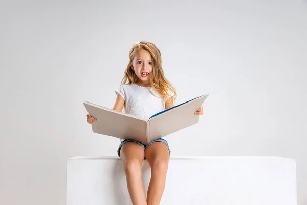 Portret małej uroczej dziewczynki w luźnych ubraniach, siedzącej na dużym pudełku, odizolowanej na białym tle studia. Szczęśliwego dzieciństwa. — Zdjęcie stockowe