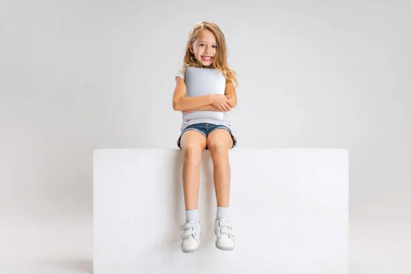 Portret małej uroczej dziewczynki w luźnych ubraniach, siedzącej na dużym pudełku, odizolowanej na białym tle studia. Szczęśliwego dzieciństwa. — Zdjęcie stockowe