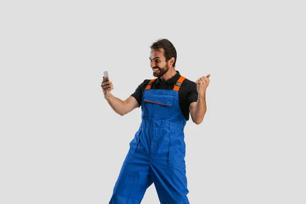 One young excited man, male auto mechanic or fitter in dungarees using phone isolated over white studio background. Concept of occupation, career, funny meme emotions — Stock Photo, Image