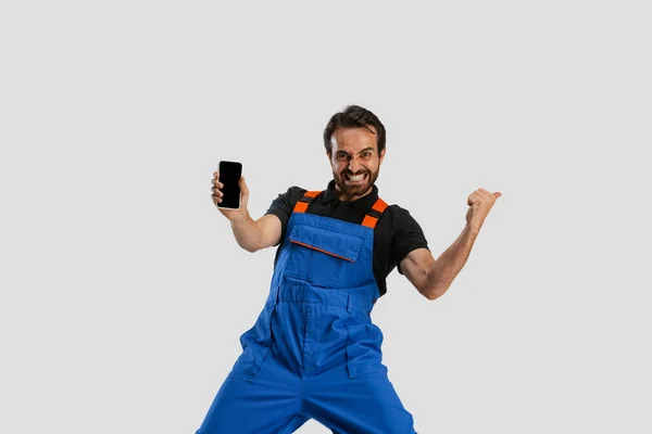 Ein junger aufgeregter Mann, männlicher Automechaniker oder Installateur in Latzhosen, der sein Telefon isoliert vor weißem Studiohintergrund benutzt. Berufsbegriff, Karriere, lustige Meme — Stockfoto