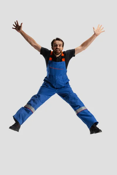 Comic portrait of young man, male auto mechanic in dungarees jumping, flying up isolated over white studio background. Concept of occupation, funny meme emotions — Stock Photo, Image